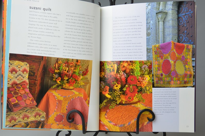 May include: A book spread showcasing a Suzani quilt and floral displays. The quilt, with its patchwork design, features a variety of shapes and colours. The flowers are arranged in a vase on a table covered with an orange tablecloth.