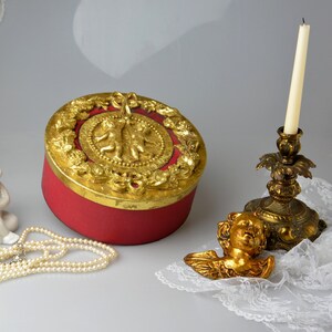 May include: A red round box with a gold lid decorated with cherubs and floral motifs. The box is sitting on a white surface next to a gold cherub figurine and a gold candlestick with a white candle.