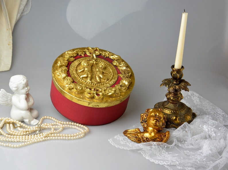 May include: A red round box with a gold lid decorated with cherubs and floral motifs. The box is sitting on a white surface next to a gold cherub figurine and a gold candlestick with a white candle.