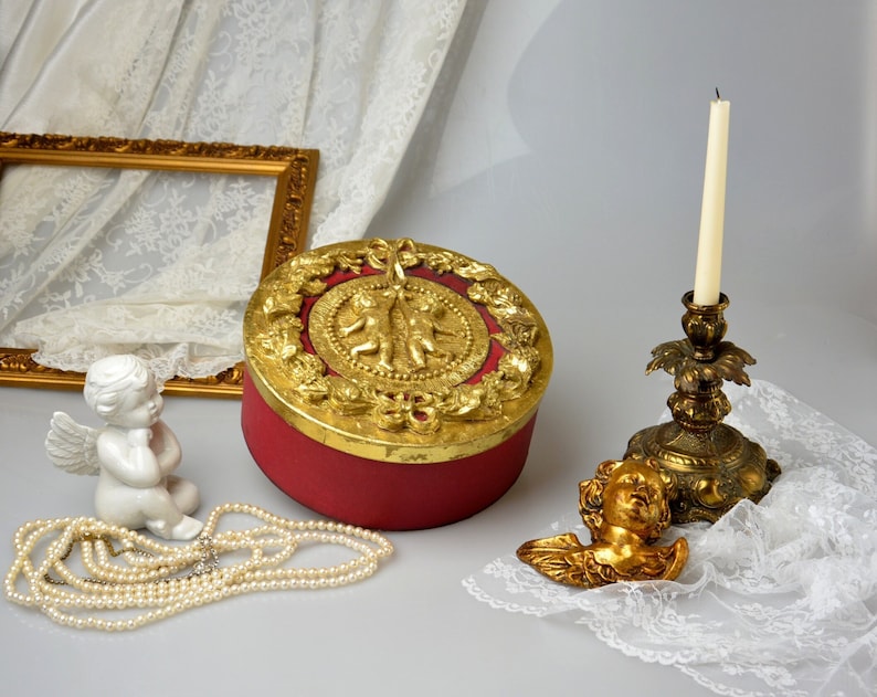 May include: A red round box with gold cherub accents and a gold trim. The box is sitting on a white lace tablecloth with a white porcelain cherub figurine, a strand of pearls, and a gold candle holder with a white candle.