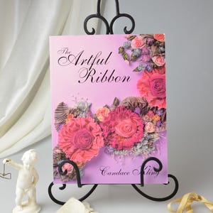 Peut inclure: Un livre intitul&eacute; "The Artful Ribbon" de Candace Kling. La couverture pr&eacute;sente un fond d&eacute;grad&eacute; rose et violet avec un arrangement floral en ruban. Les fleurs sont roses, blanches et violettes.
