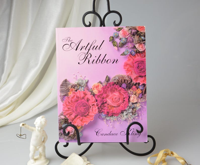 Peut inclure: Un livre intitul&eacute; "The Artful Ribbon" de Candace Kling. La couverture pr&eacute;sente un fond d&eacute;grad&eacute; rose et violet avec un arrangement floral en ruban. Les fleurs sont roses, blanches et violettes.