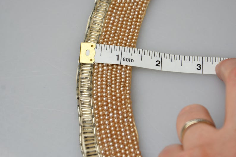 May include: A close-up of a pearl necklace with a gold-toned metal clasp. The necklace is being measured with a tape measure, showing it is 1 1/2 inches long.