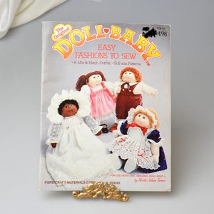 May include: A vintage "Doll Baby" sewing pattern booklet featuring illustrations of dolls in various outfits. The cover has the text "Easy Fashions to Sew" and "8 Mix & Match Outfits". The booklet is held by a gold-colored decorative clip.