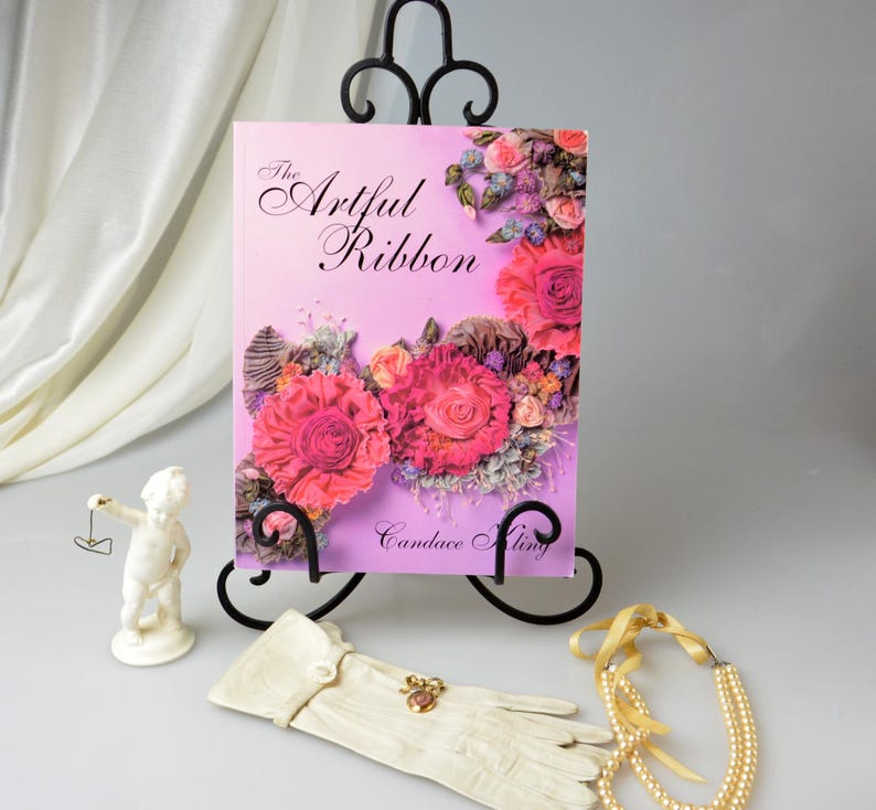 May include: A book titled 'The Artful Ribbon' by Candace Kling. The book cover features a pink background with a floral design made of ribbon. The book is displayed on a black metal book stand.
