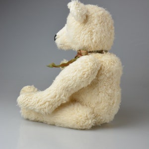 May include: A white plush teddy bear sitting with its legs crossed. The bear has a green and brown ribbon around its neck.