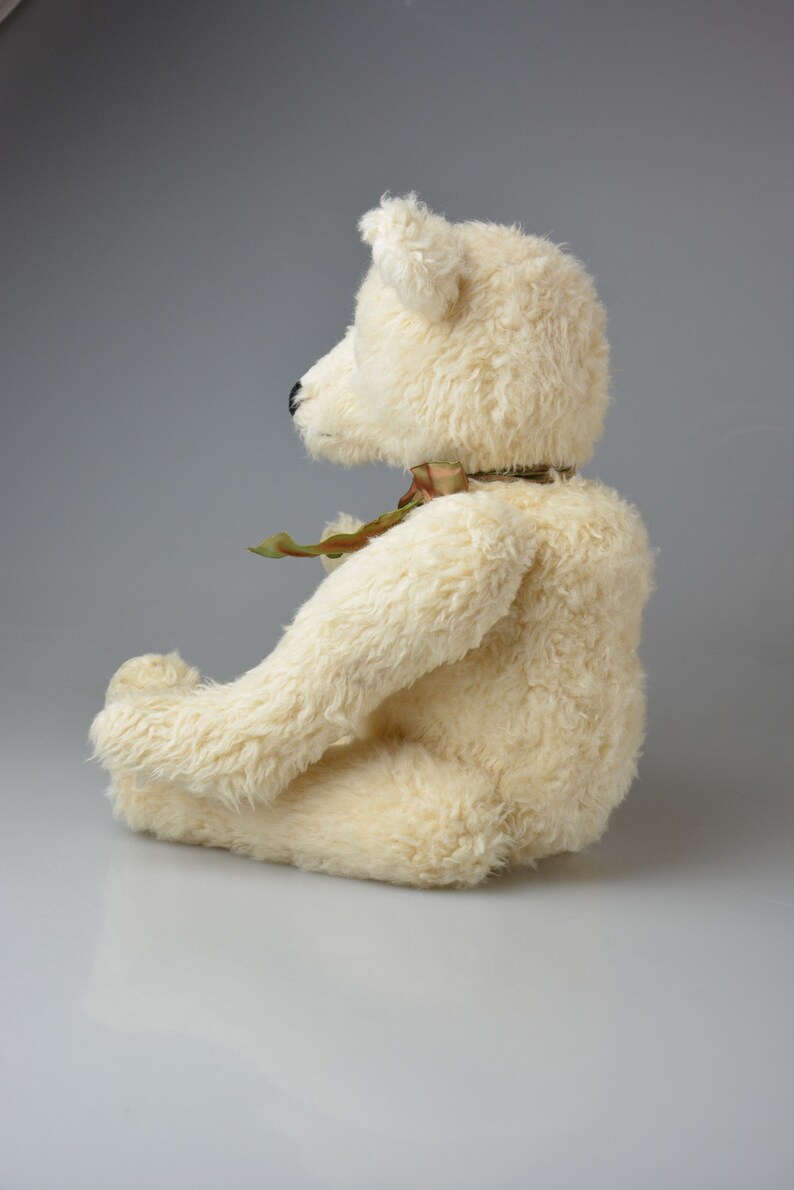 May include: A white plush teddy bear sitting with its legs crossed. The bear has a green and brown ribbon around its neck.