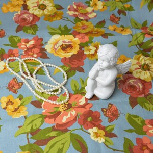 Może przedstawiać: Biała porcelanowa figurka anioła z skrzydłami, siedząca na niebieskim materiale z kwiatowym wzorem. Na materiale leży naszyjnik z pereł.