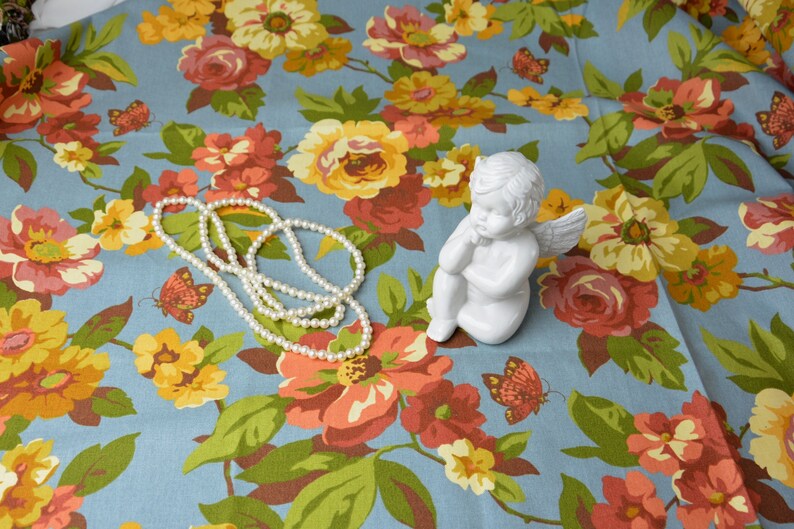 Może przedstawiać: Biała porcelanowa figurka anioła z skrzydłami, siedząca na niebieskim materiale z kwiatowym wzorem. Na materiale leży naszyjnik z pereł.