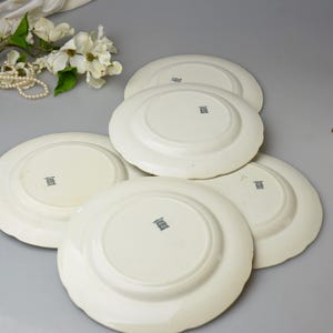 May include: A collection of off-white ceramic plates with scalloped edges. The plates are stacked, revealing their undersides, each with a small, dark emblem. A pearl necklace and white flowers are in the background.