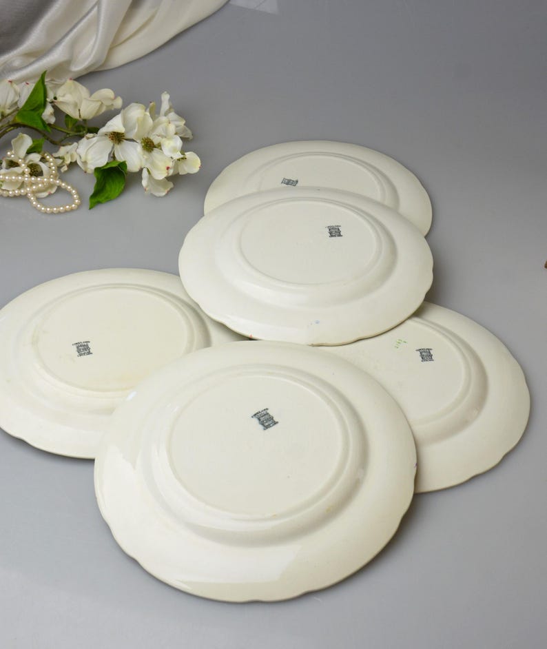 May include: A collection of off-white ceramic plates with scalloped edges. The plates are stacked, revealing their undersides, each with a small, dark emblem. A pearl necklace and white flowers are in the background.