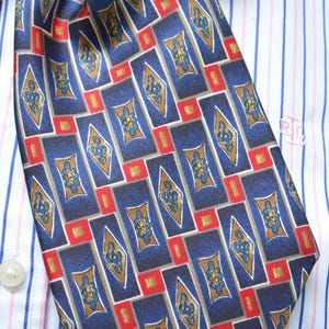 May include: A necktie featuring a repeating pattern of dark blue rectangles, gold diamond shapes, and small red squares, worn over a white dress shirt with thin blue and red vertical stripes and a small embroidered logo on the chest area.