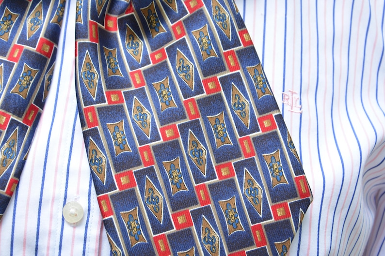 May include: A necktie featuring a repeating pattern of dark blue rectangles, gold diamond shapes, and small red squares, worn over a white dress shirt with thin blue and red vertical stripes and a small embroidered logo on the chest area.