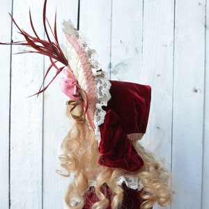 May include: A red velvet bonnet with a white and pink lace trim and a red feather accent.