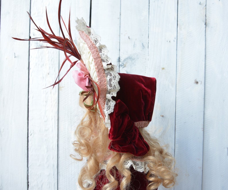 May include: A red velvet bonnet with a white and pink lace trim and a red feather accent.