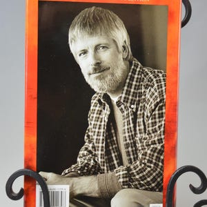 May include: A book cover with a portrait of a man in a plaid shirt, displayed in a decorative black metal stand. The book cover has an orange border and the text "A CROSSINGS BOOK CLUB EDITION". The man has gray hair and a beard.