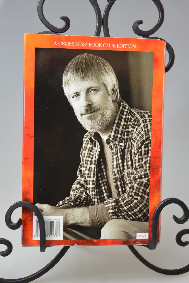 May include: A book cover with a portrait of a man in a plaid shirt, displayed in a decorative black metal stand. The book cover has an orange border and the text "A CROSSINGS BOOK CLUB EDITION". The man has gray hair and a beard.