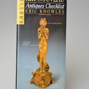 May include: A gold-coloured Art Nouveau statue of a woman holding flowers, displayed with a book titled "Art Nouveau Antiques Checklist" by Eric Knowles. The book is propped up by a gold-coloured stand.