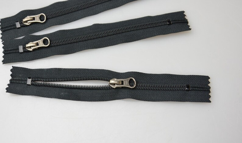 May include: Three black zipper closures with silver pull tabs. The zippers are laid out on a white surface.