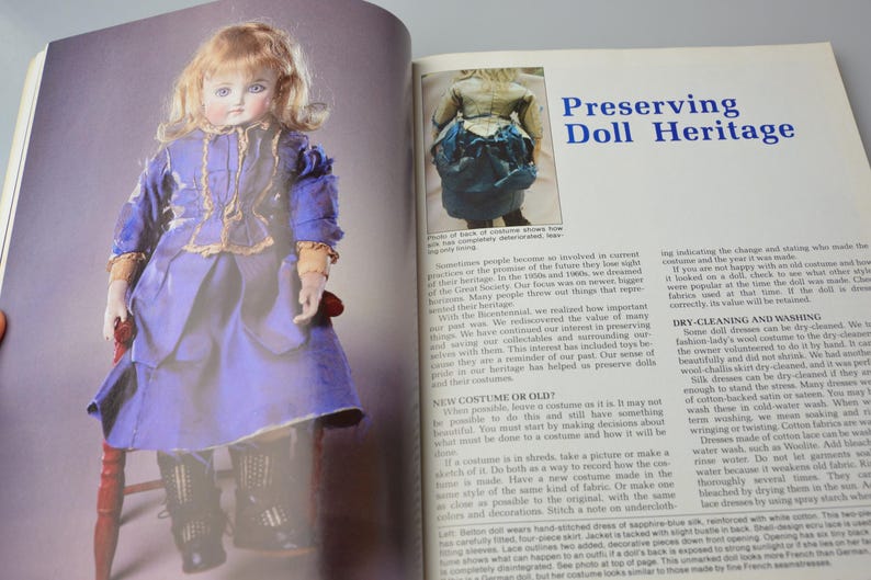 Pode incluir: Uma boneca de porcelana vintage com um vestido azul e dourado est&aacute; sentada em uma cadeira de madeira. A boneca tem um rosto delicado com olhos azuis pintados e bochechas coradas. A imagem &eacute; de um livro sobre a preserva&ccedil;&atilde;o do patrim&ocirc;nio das bonecas.