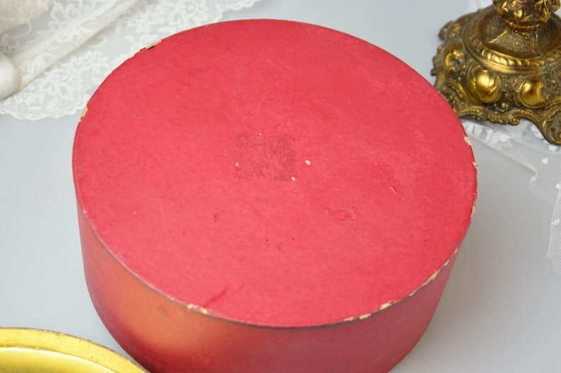 May include: A round, red cardboard box with a smooth, slightly worn surface. The box is empty and appears to be vintage.