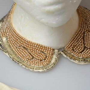May include: A gold and cream beaded collar necklace with a scalloped edge. The necklace is made of small, round beads that are strung together in a delicate pattern.