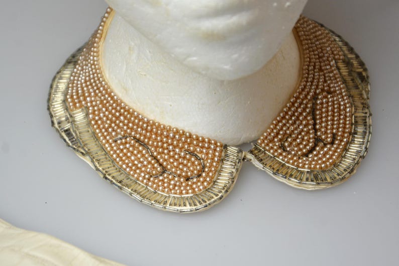 May include: A gold and cream beaded collar necklace with a scalloped edge. The necklace is made of small, round beads that are strung together in a delicate pattern.