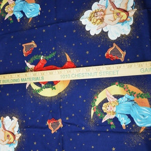 May include: Dark blue fabric with a repeating pattern of cherubs, harps, moons, and stars. The cherubs are depicted in various poses, some playing instruments. The fabric has a celestial theme, with gold accents and a wooden ruler across the image.