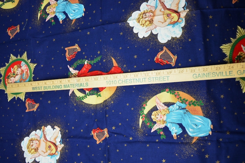 May include: Dark blue fabric with a repeating pattern of cherubs, harps, moons, and stars. The cherubs are depicted in various poses, some playing instruments. The fabric has a celestial theme, with gold accents and a wooden ruler across the image.