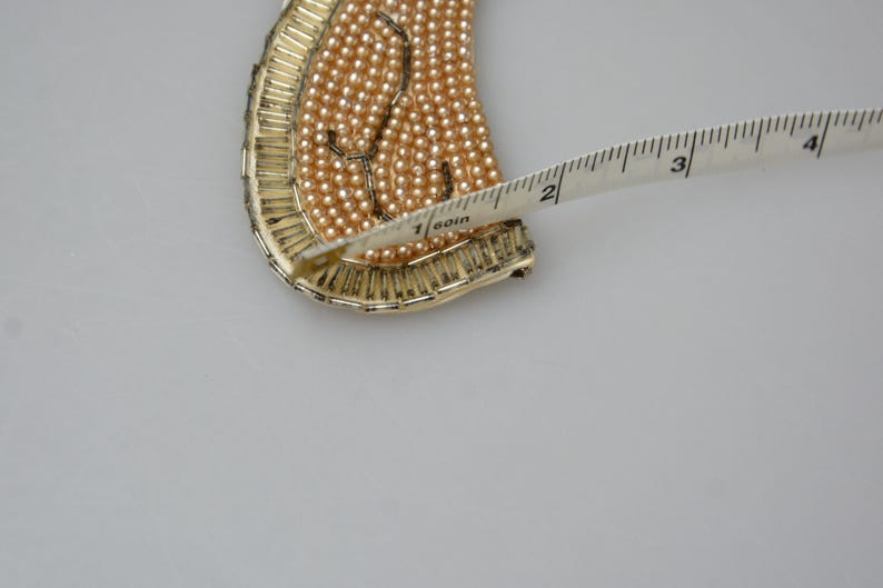May include: A gold-toned metal necklace with a design of small, light brown beads. The necklace is being measured with a tape measure, showing a length of 1 1/2 inches.