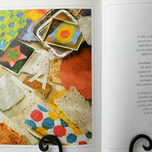 May include: A close-up of a book page with the title "Papermaking". The page features a colorful collage of handmade paper samples, including a star-shaped piece, a letter "A", and a tray of colorful paper. The text on the page describes the process of making handmade paper.