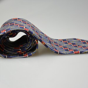 May include: A necktie featuring a repeating geometric pattern of navy blue rectangles with gold-toned emblem accents and red border details. The fabric has a smooth, satin-like finish, and the tie is partially coiled to display the intricate design.