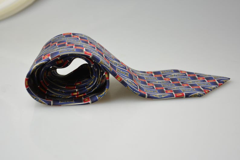 May include: A necktie featuring a repeating geometric pattern of navy blue rectangles with gold-toned emblem accents and red border details. The fabric has a smooth, satin-like finish, and the tie is partially coiled to display the intricate design.