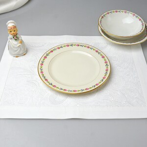 May include: A white placemat with a floral design, a decorative figurine, and a set of fine china. The china includes a dinner plate and two smaller bowls, all with a gold rim and a pink and blue floral border.