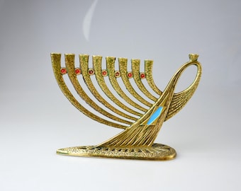 Puede incluir: Una menorá dorada con nueve candelabros, cada uno adornado con una joya roja. La menorá presenta un diseño abstracto con un acento azul y está sobre un fondo blanco.
