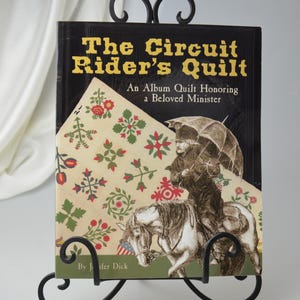 Op de afbeelding: Een zwart boek getiteld "The Circuit Rider's Quilt: An Album Quilt Honoring a Beloved Minister" door Jennifer Dick. De omslag van het boek toont een kleurrijk quiltpatroon en een zwart-witte illustratie van een man die op een paard rijdt.