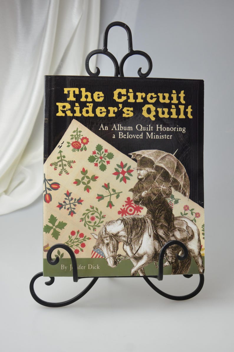 Może przedstawiać: Czarna książka zatytułowana "The Circuit Rider's Quilt: An Album Quilt Honoring a Beloved Minister" autorstwa Jennifer Dick. Okładka książki przedstawia kolorowy wz&oacute;r patchworku i czarno-białą ilustrację mężczyzny jadącego konno.