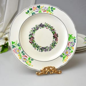 May include: A white ceramic plate with a floral wreath design in the center and a floral border. The plate has a scalloped edge and a black trim. The plate is displayed on a gold-colored stand.