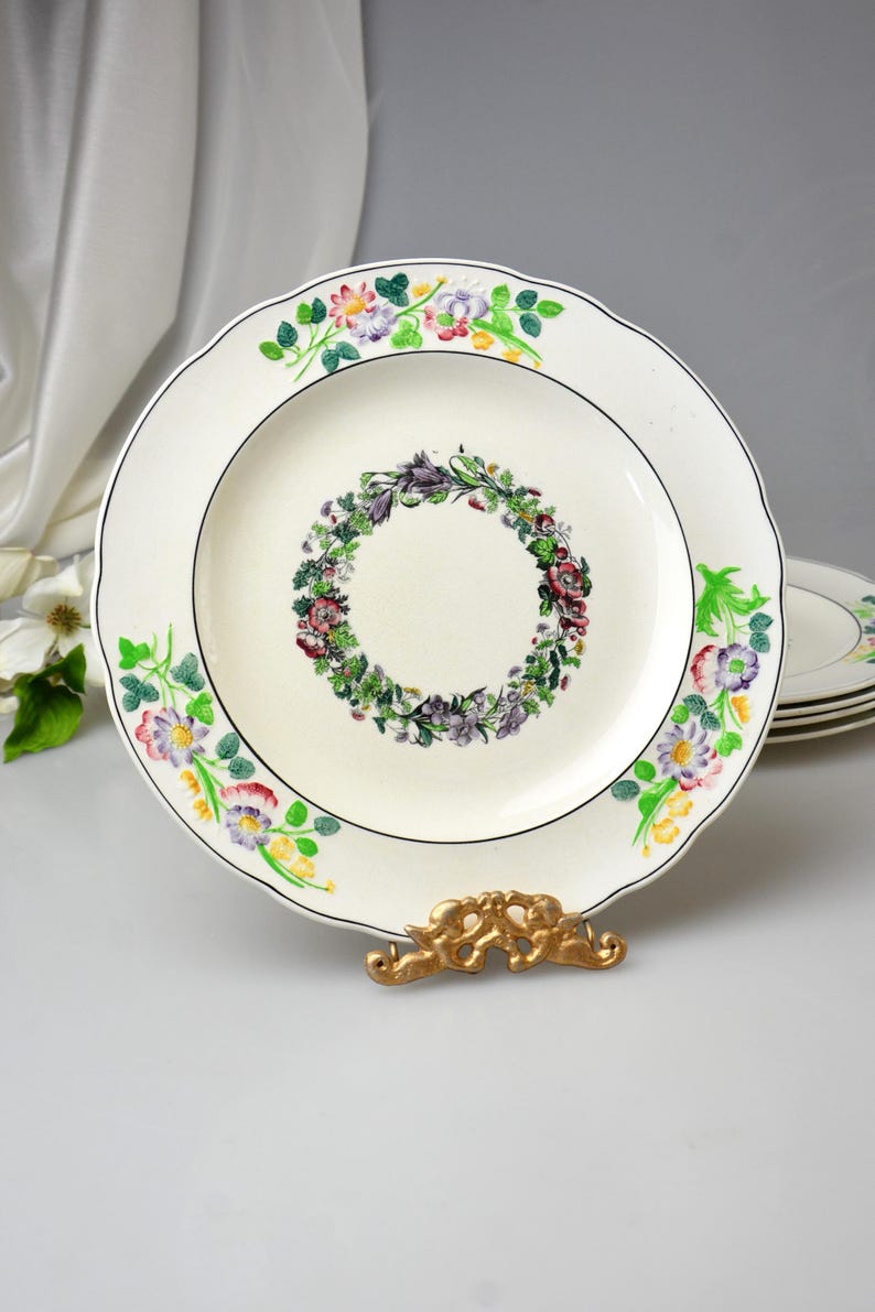 May include: A white ceramic plate with a floral wreath design in the center and a floral border. The plate has a scalloped edge and a black trim. The plate is displayed on a gold-colored stand.