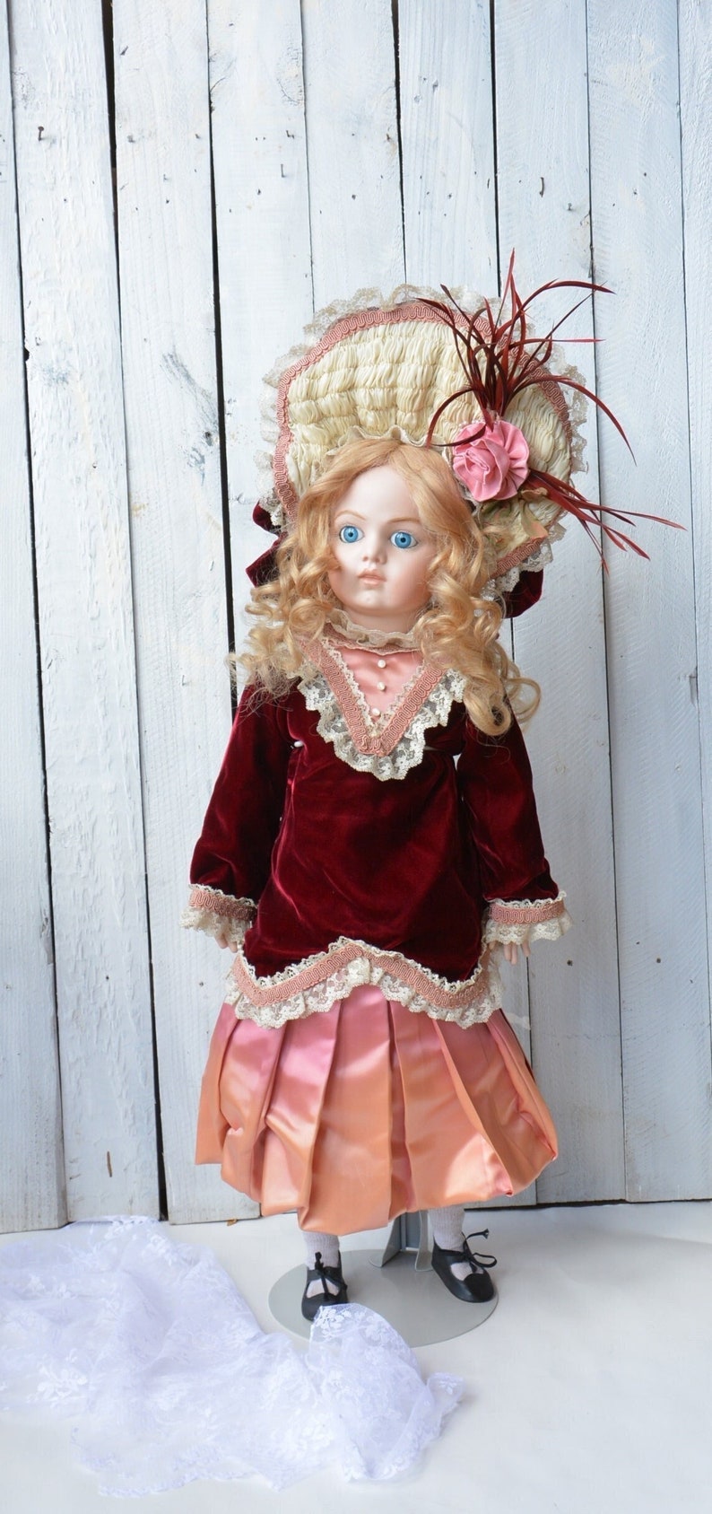 May include: A porcelain doll wearing a red velvet dress with a lace trim, a pink satin skirt, and a white hat with a pink flower. The doll has blonde hair and blue eyes.