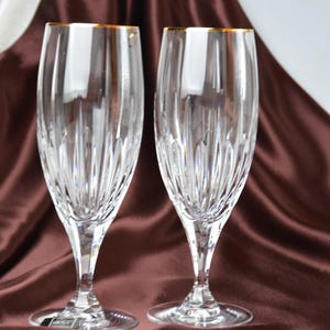 Pu&ograve; includere: Due fl&ucirc;te da champagne in cristallo trasparente con bordi dorati. I bicchieri presentano un motivo verticale strutturato e sono esposti su un tessuto di raso marrone. Le fl&ucirc;te sono eleganti e adatte alle occasioni speciali.
