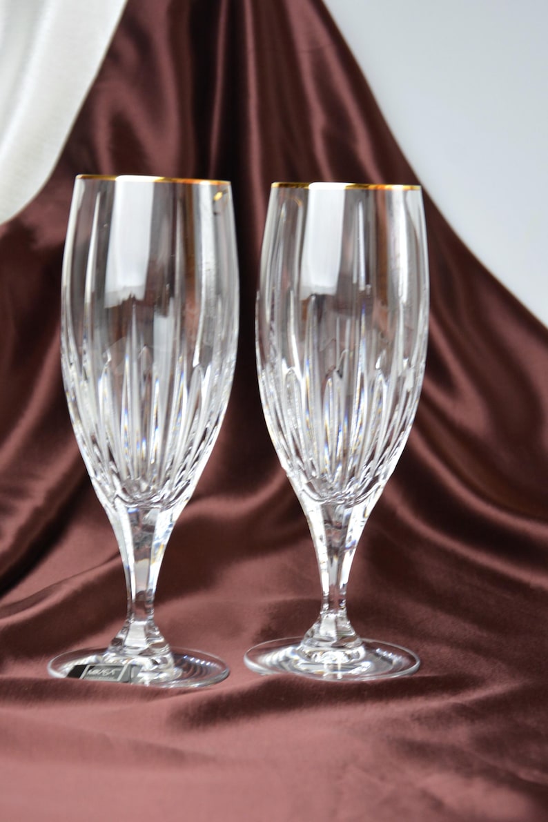 Pu&ograve; includere: Due fl&ucirc;te da champagne in cristallo trasparente con bordi dorati. I bicchieri presentano un motivo verticale strutturato e sono esposti su un tessuto di raso marrone. Le fl&ucirc;te sono eleganti e adatte alle occasioni speciali.