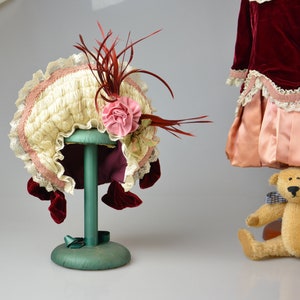 May include: A white and pink doll bonnet with a red feather trim and a pink flower accent. The bonnet is displayed on a green stand.