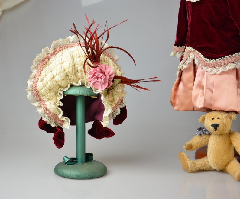 May include: A white and pink doll bonnet with a red feather trim and a pink flower accent. The bonnet is displayed on a green stand.