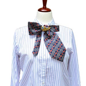 May include: A white long-sleeved shirt with thin blue and red vertical stripes, featuring a dark blue necktie with a repeating geometric pattern of red and gold diamonds. A gold-toned feather brooch with a blue stone center secures the tie.