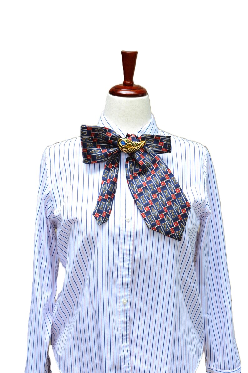 May include: A white long-sleeved shirt with thin blue and red vertical stripes, featuring a dark blue necktie with a repeating geometric pattern of red and gold diamonds. A gold-toned feather brooch with a blue stone center secures the tie.