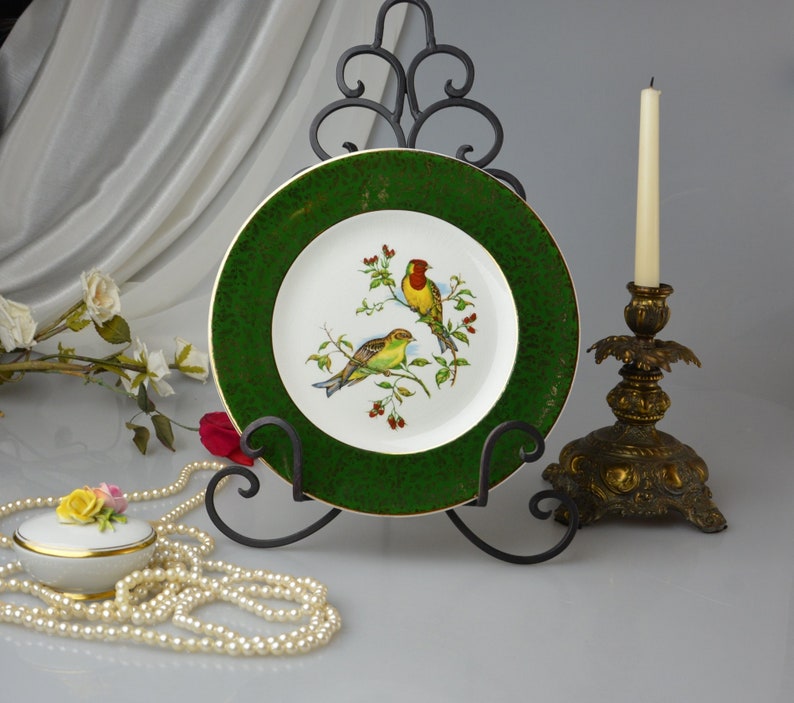 May include: A white porcelain plate with a green rim and a gold flecked design. The plate features a painted image of two birds on a branch with red berries. The plate is displayed on a black metal stand.