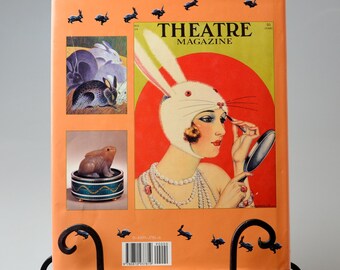 May include: The back cover of a Theatre Magazine from 1935. The cover features a woman wearing a white rabbit costume and applying makeup. The magazine cover is orange with a red circle in the center. The magazine is on a black metal stand.