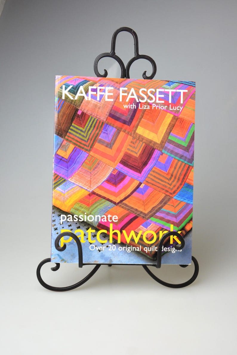May include: A book titled "KAFFE FASSETT" with "passionate patchwork" on the cover, displayed on a black metal stand. The book cover features a colourful patchwork design in shades of orange, purple, and green. The stand has decorative scrollwork.