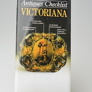 May include: A book titled "MILLER'S Antiques Checklist VICTORIANA" is displayed on a gold-colored stand. The book cover features a black background with gold lettering and an image of a decorative plate with a parrot and cat.